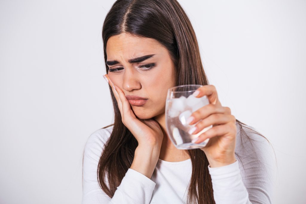 Mujer con expresión de dolor dental, sosteniendo su mejilla mientras tiene un vaso de agua con hielo